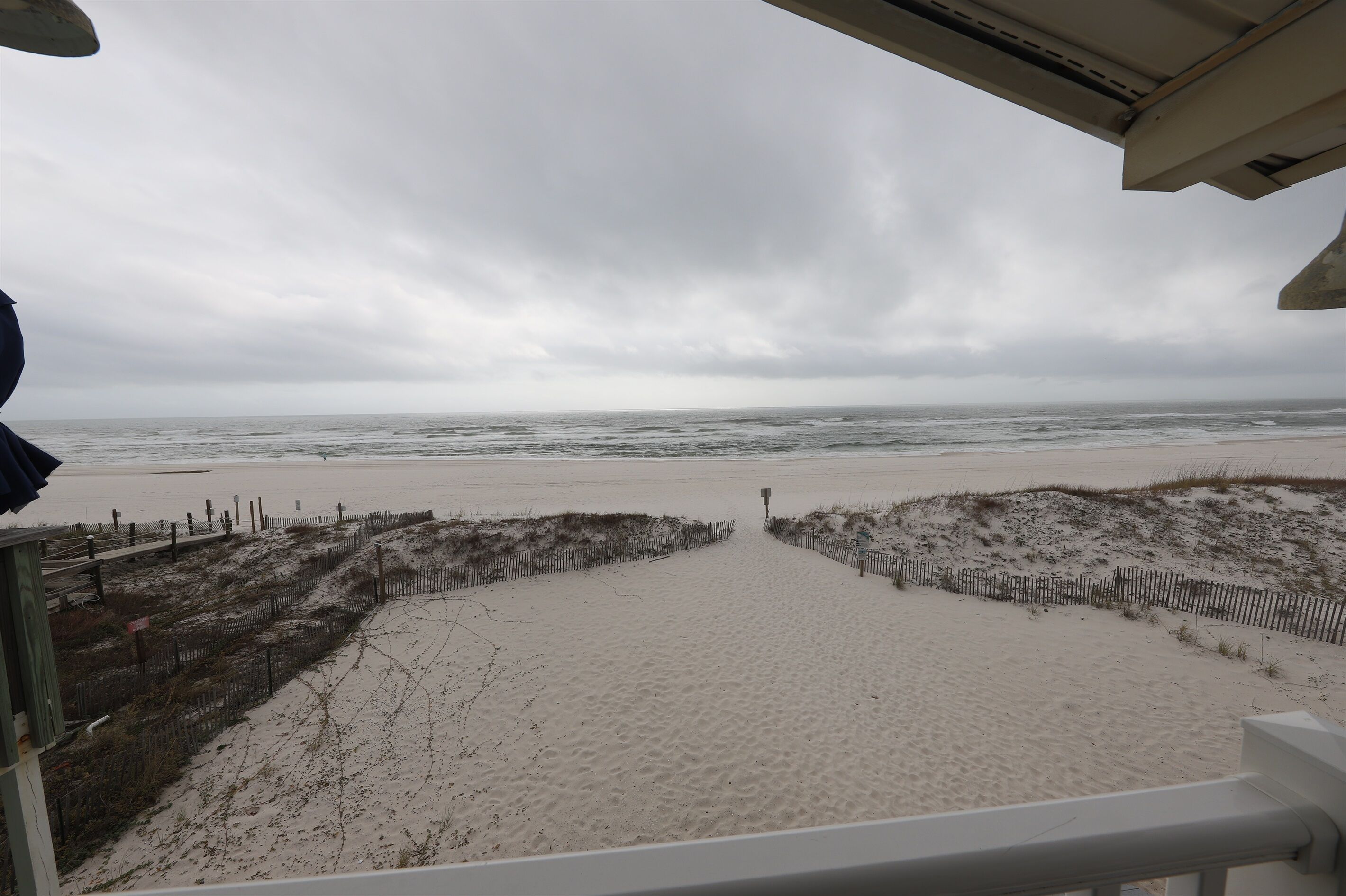 A beach view with a sandy path leading to the water, surrounded by dunes and a wooden fence.