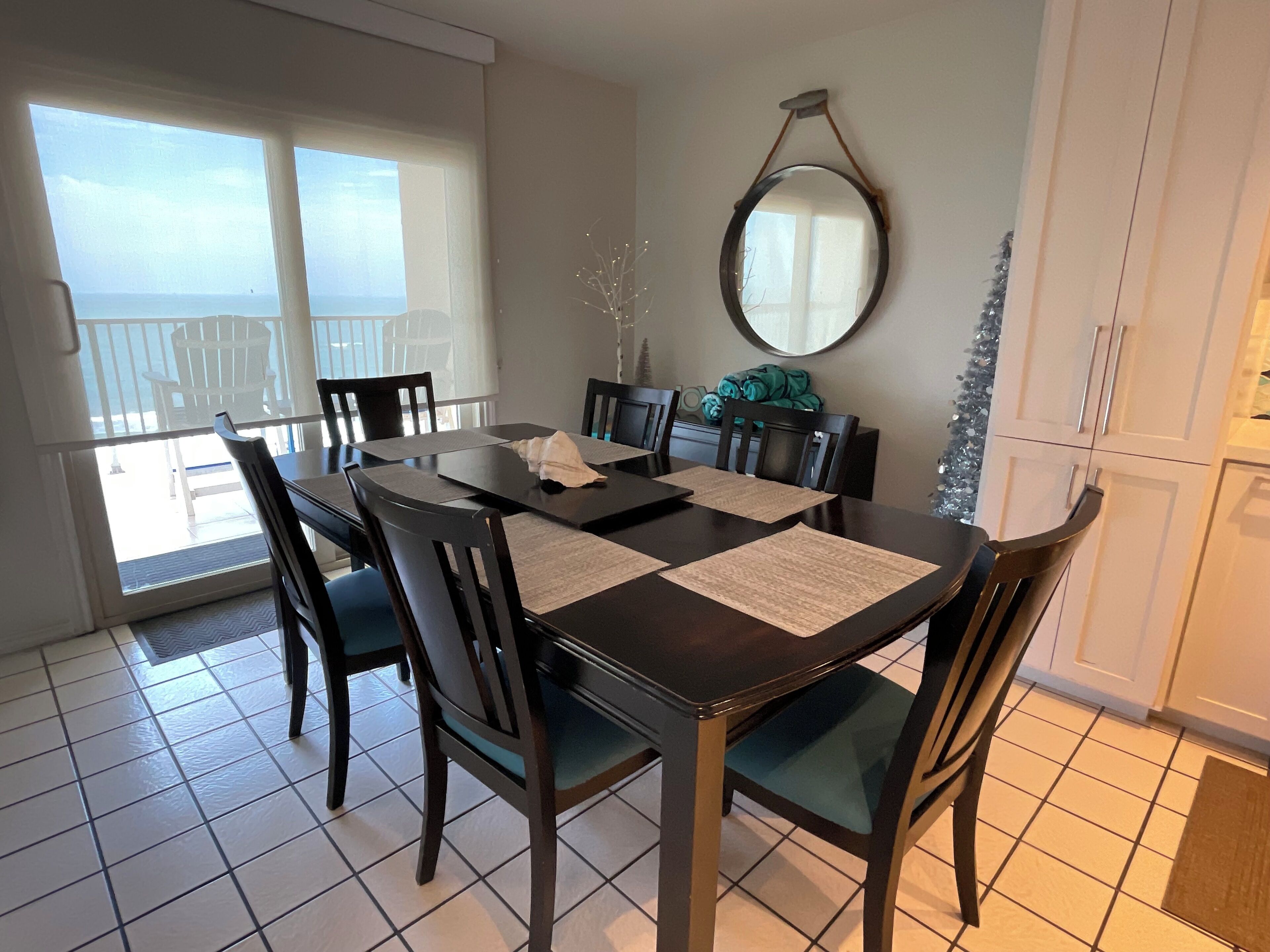 A dining area with a dark wooden table and chairs, a large mirror on the wall, and a view of the sea through the windows.