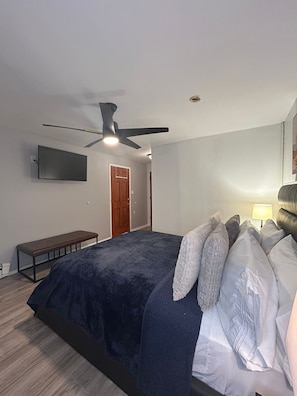 Une chambre à coucher comprenant un lit, un ventilateur de plafond, un téléviseur fixé au mur, un banc et une porte.