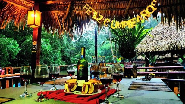 Un espace de restauration en plein air avec des tables à toit de chaume, des verres à vin et des bouteilles, le tout entouré d’une végétation luxuriante.