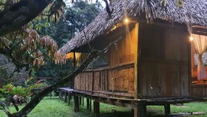 Une construction rustique en bois, dotée d’un toit de chaume et surélevée sur des piliers, avec une petite véranda comprenant des chaises et une table.