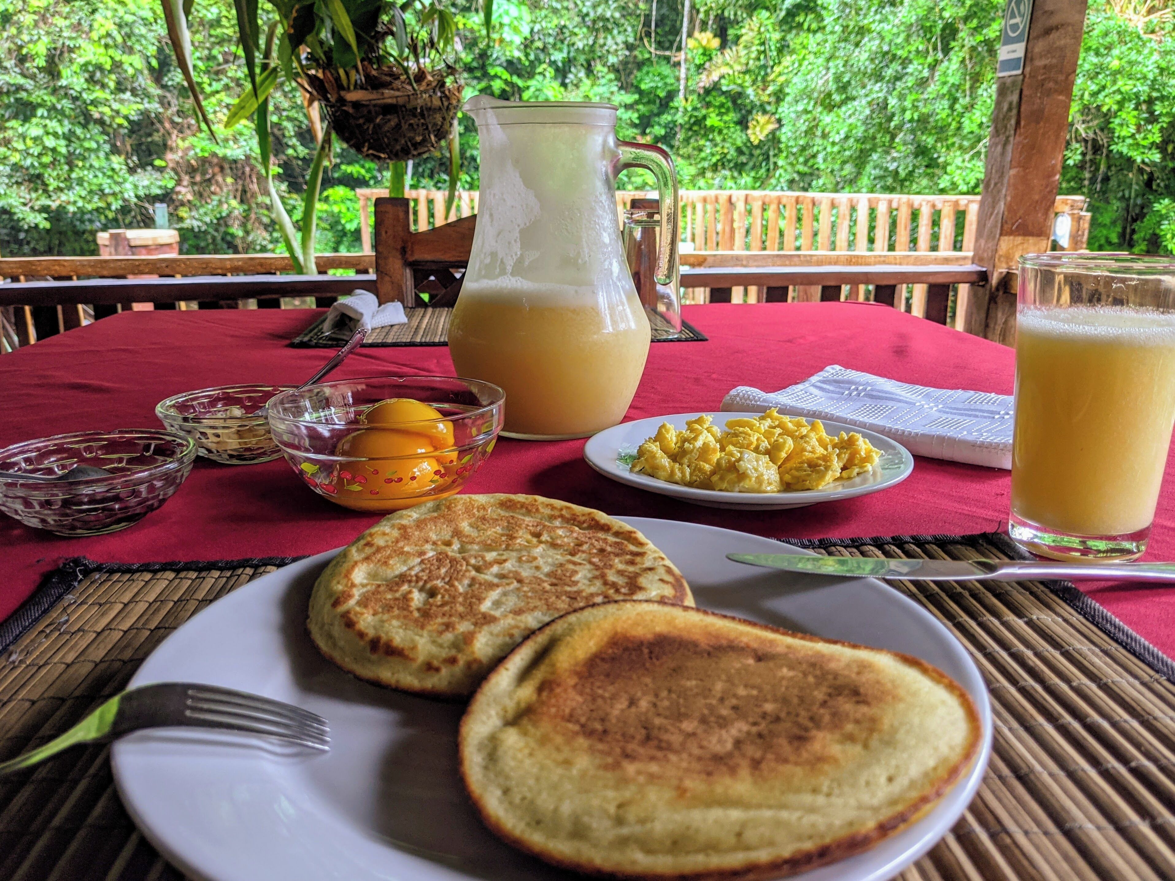Desayuno con tortitas, huevos revueltos y zumo de naranja sobre un mantel rojo.
