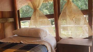 Una habitación de madera con una cama, una mesita y un ventanal con cortina mosquitera.