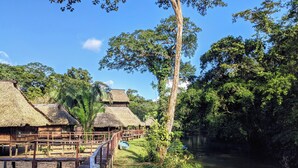 Une terrasse en bois menant à des huttes aux toits de chaume, situées au bord d’une rivière et entourées d’une végétation luxuriante.