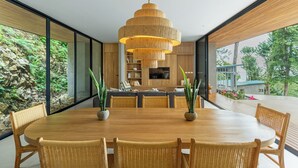 A modern dining area with a wooden table and wicker chairs, a multi-tiered wooden pendant light, and a view of a rocky garden through large windows. - Casa Roca: Luxury Jungle Retreat with Natural Design & Infinity Pool (Santa Teresa de Cobano)