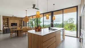 A modern kitchen with a central island, wooden cabinetry, and large windows offering a view of the outdoors. - Casa Roca: Luxury Jungle Retreat with Natural Design & Infinity Pool (Santa Teresa de Cobano)