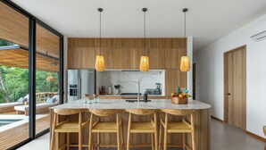 A modern kitchen with a central island, wooden bar stools, pendant lights, and a view of the outdoors. - Casa Roca: Luxury Jungle Retreat with Natural Design & Infinity Pool (Santa Teresa de Cobano)