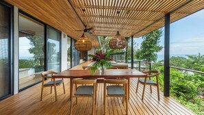 A wooden deck with a table and chairs, large windows, and a view of the outdoors. - Casa Roca: Luxury Jungle Retreat with Natural Design & Infinity Pool (Santa Teresa de Cobano)
