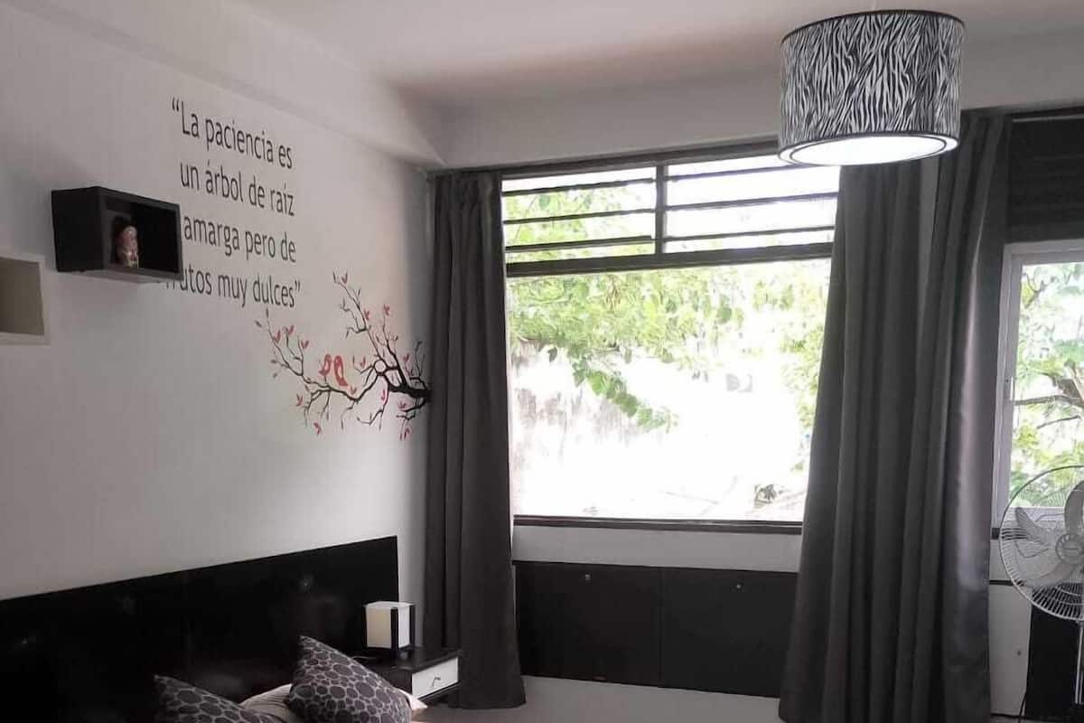 Ein modernes Hotelzimmer mit einem großen Bett, einem Nachttisch, einem Wandregal, einem Fenster mit Vorhängen und einem Deckenventilator.