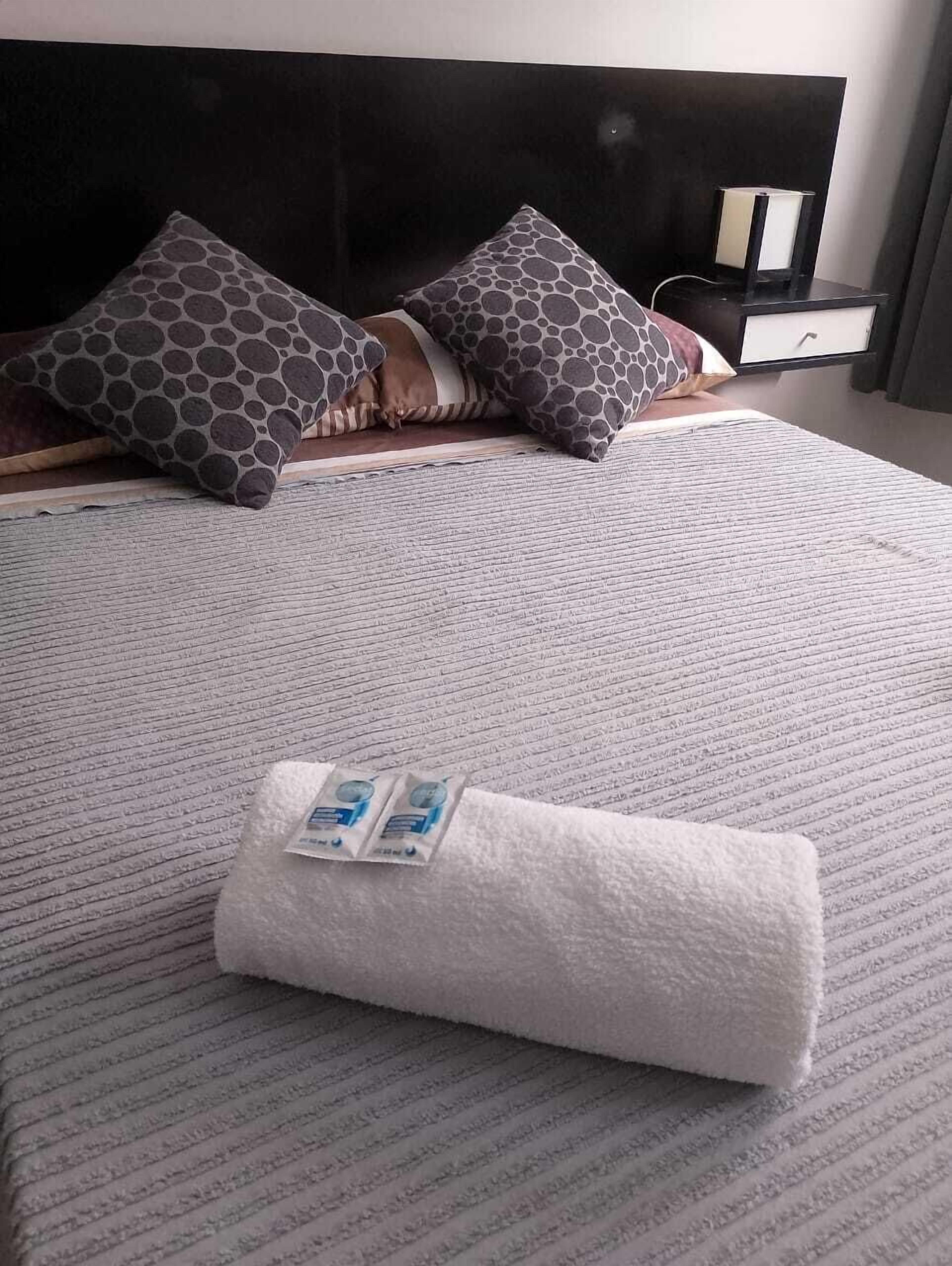 A neatly made bed with patterned pillows, a folded towel, and a bedside table with a lamp.