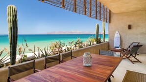 Une table en bois, un bocal en verre, une planche de surf et une chaise sont disposés sur un balcon donnant sur une plage, avec un cactus et un paysage désertique en toile de fond.