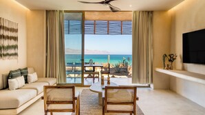 Un salon comprenant un canapé, deux fauteuils, une table et un ventilateur de plafond. Une grande fenêtre offre une vue sur l’océan et les montagnes.
