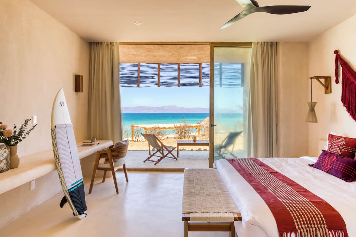 Ett hotellrum med en säng, ett skrivbord, en stol, en surfbräda och utsikt över stranden och havet.