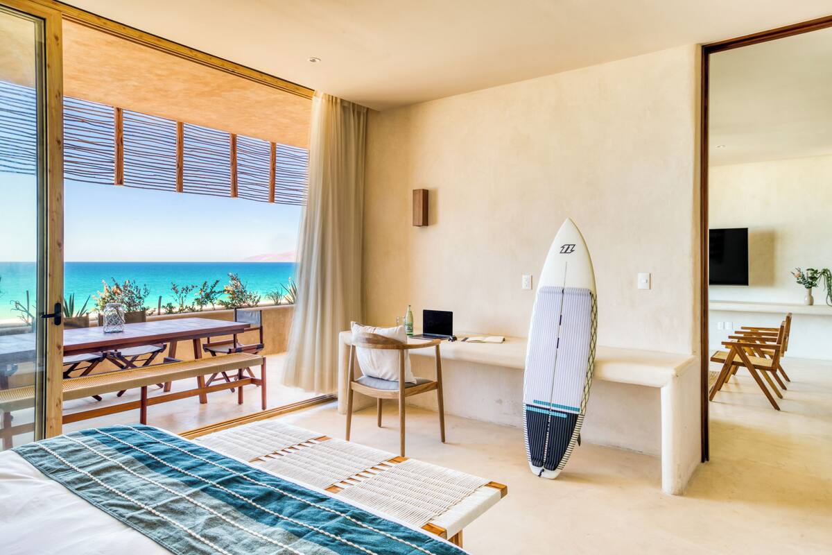 Ett hotellrum med en säng, ett skrivbord, en stol, en surfbräda och utsikt över havet.