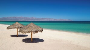 Une plage avec deux parasols en paille et une vue dégagée sur l’océan et les montagnes en arrière-plan.