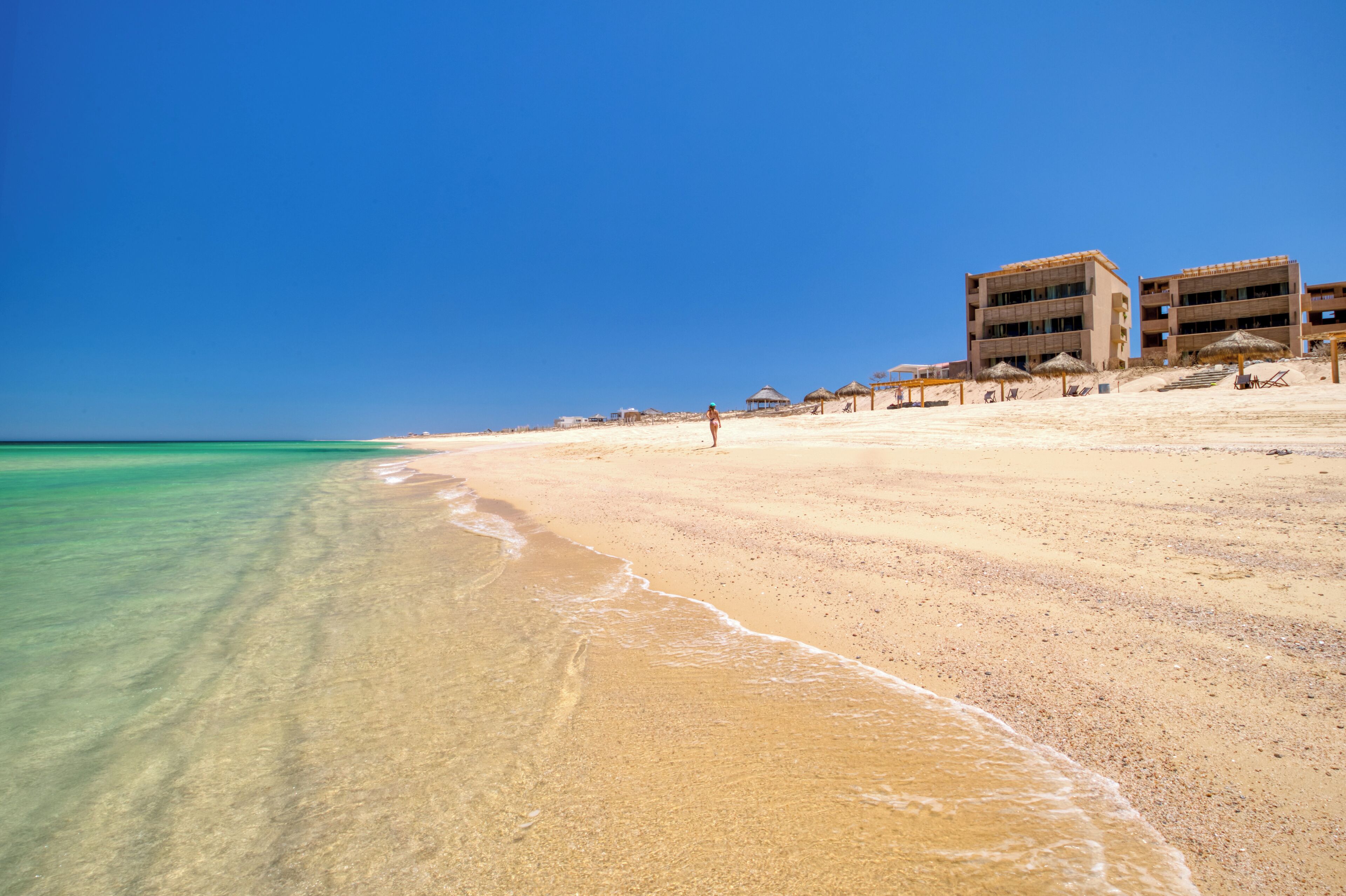 A beach with clear turquoise water, a sandy shore, and a building with multiple balconies.