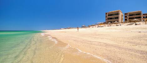 A beach with clear turquoise water, a sandy shore, and a building with multiple balconies.