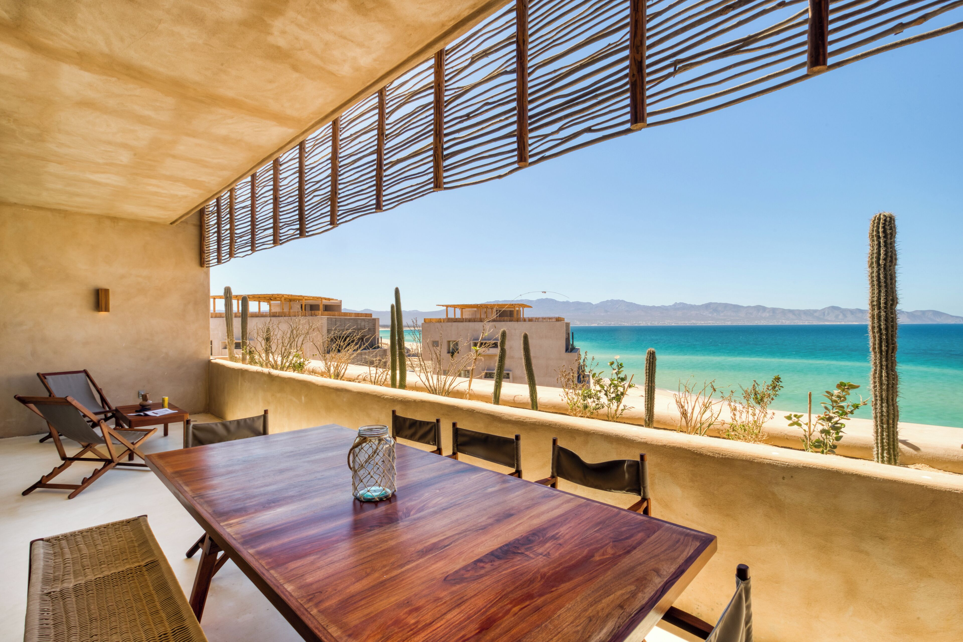 Une vaste terrasse dotée d’une table et de chaises en bois, offrant une vue sur une plage parsemée de cactus et un ciel d’un bleu limpide.