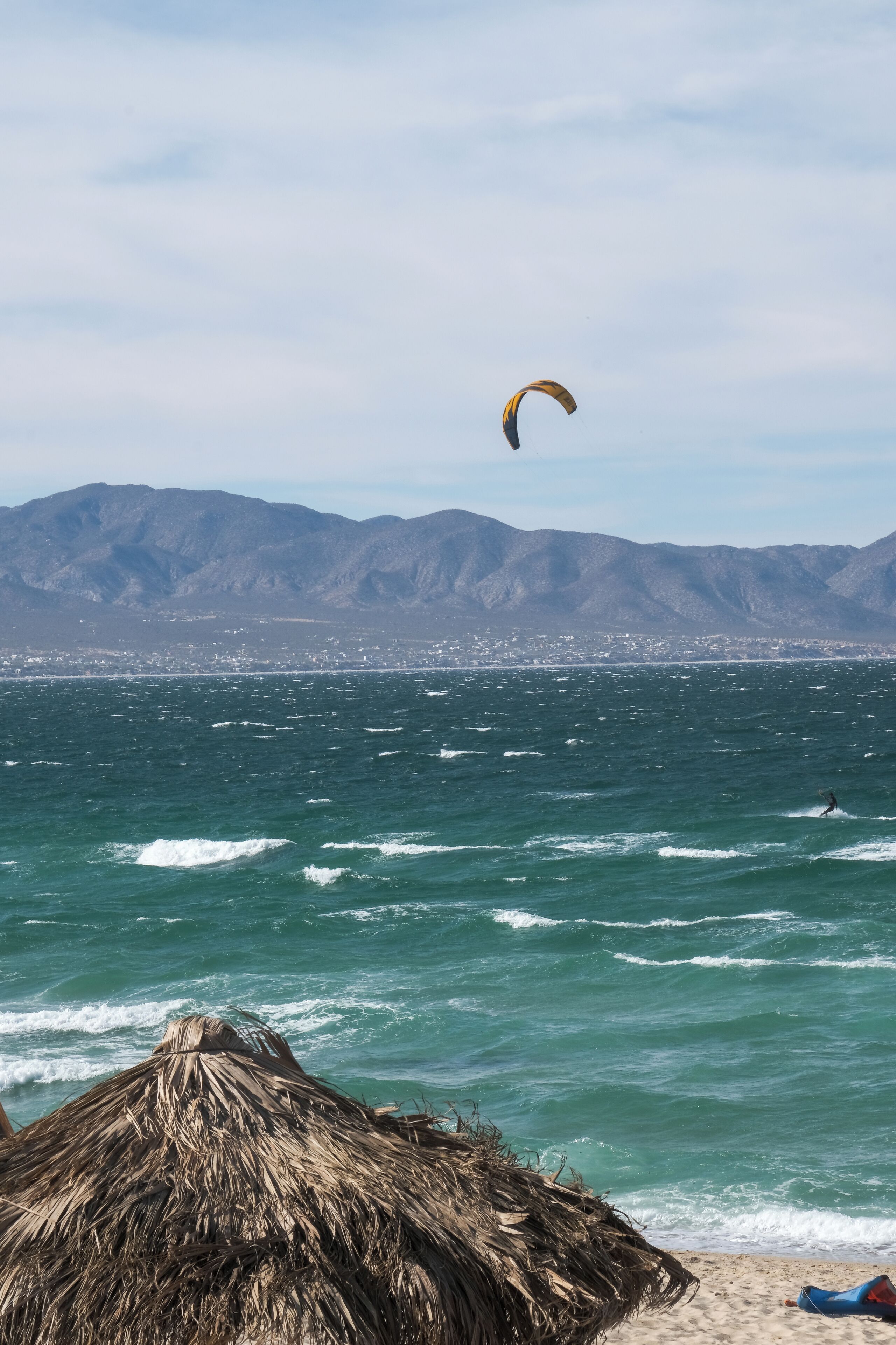 Una playa con una cabaña de techo de paja, una persona practicando kitesurf y montañas al fondo.