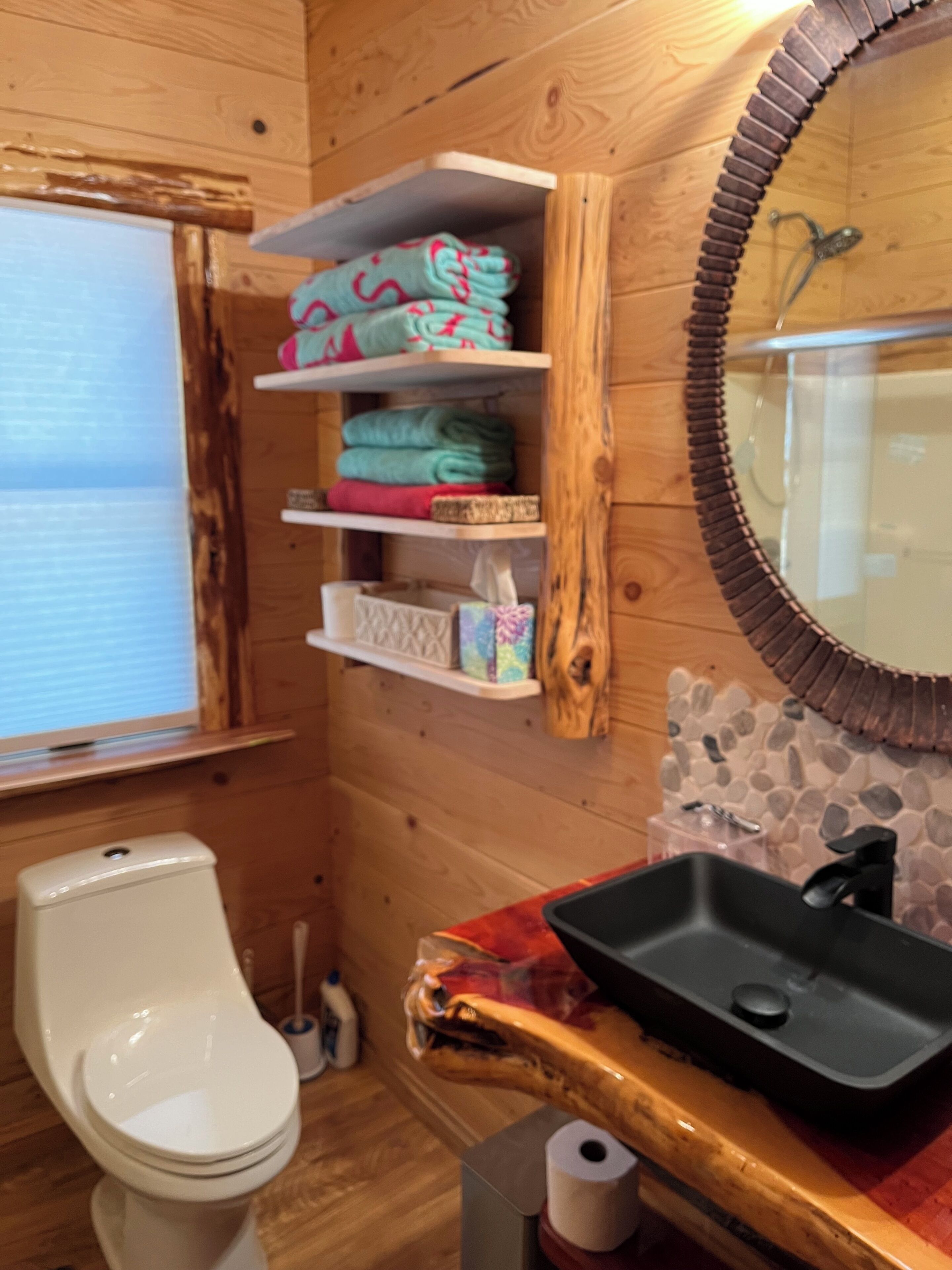 A wooden cabin bathroom with a toilet, shelves with folded towels, a sink with a black basin, and a stone backsplash.