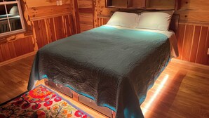 Una habitación tipo cabaña de madera, con una cama, una ventana y una estantería con objetos.