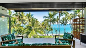 Ein Balkon mit zwei Holzstühlen und einem Tisch, der auf einen Strand mit Palmen und das Meer blickt.