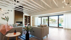 A modern living room with a sofa, a small table, and a bookshelf. There is a large window with a view of the outside. - Sunset 4BR Waterfront Villa in Golden Isles w/ Heated Pool & Spa (Hallandale Beach)