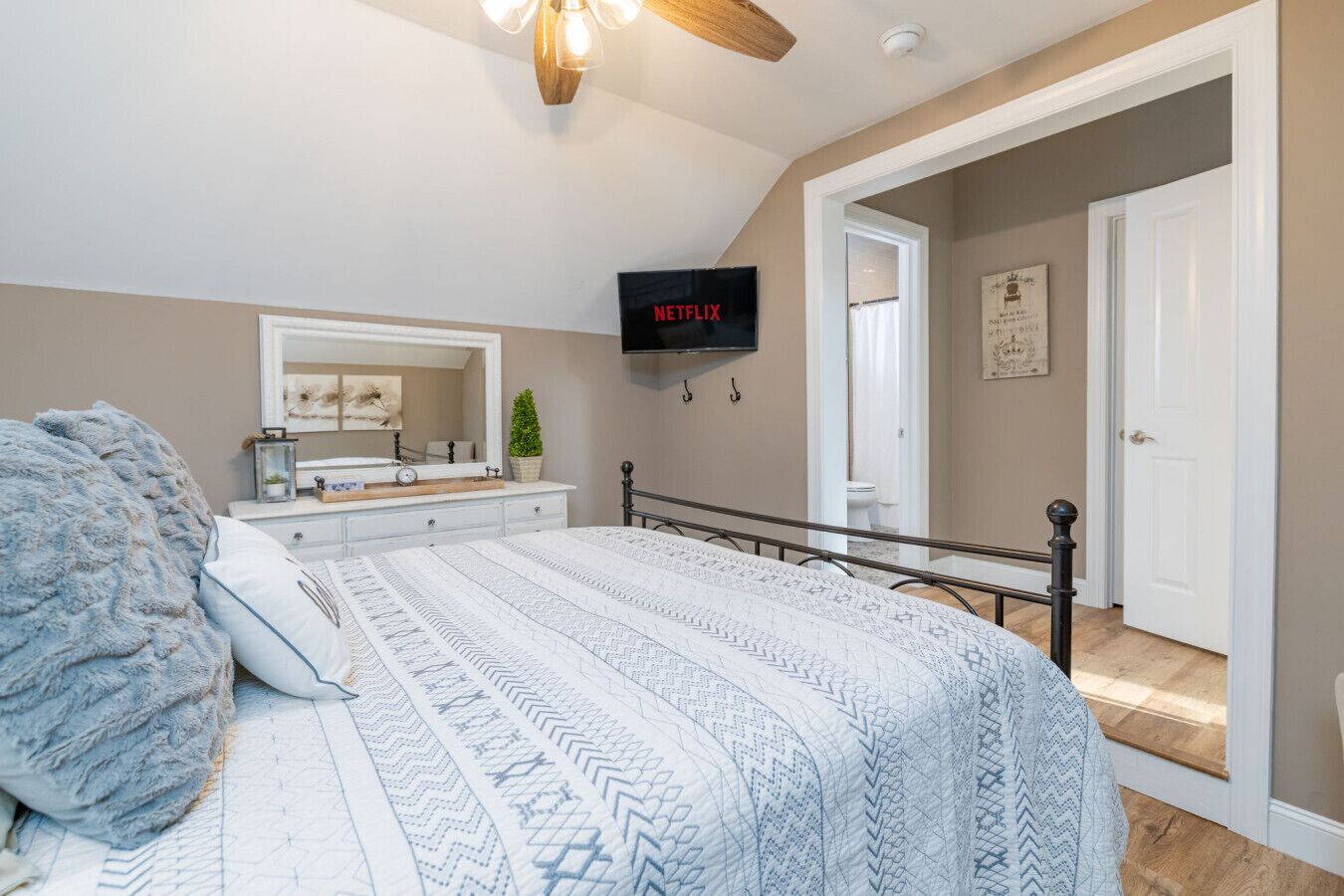 A bedroom with a bed, a dresser, a mirror, a TV mounted on the wall, and a door leading to another room.