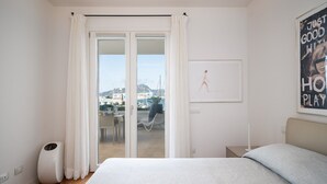 A bedroom with a large bed, a view of the city through a sliding door, a small table with a chair, and a poster on the wall. - Mobiretreat by Klodge - waterfront design flat (Olbia)