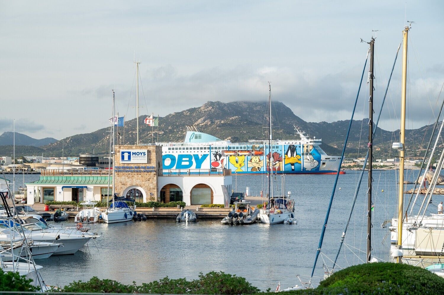 Un puerto deportivo con barcos amarrados, un barco colorido con personajes de dibujos animados y un edificio con la señalización 'MAYS' y 'OBY'.
