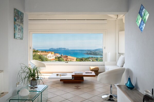 A modern living room with a large window overlooking a coastal view, a glass coffee table, and two white armchairs.