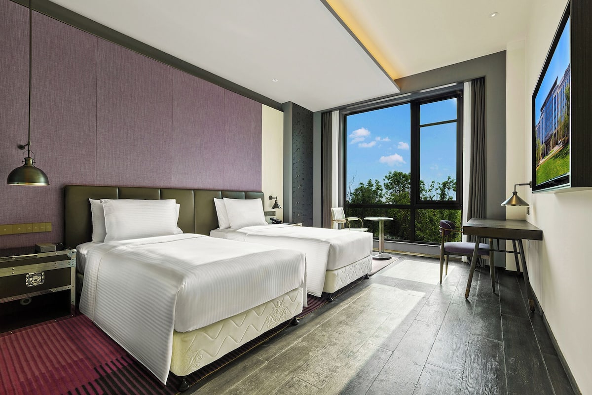 A modern hotel room with two beds, a desk, a chair, a TV, and a large window offering a view of the outdoors.