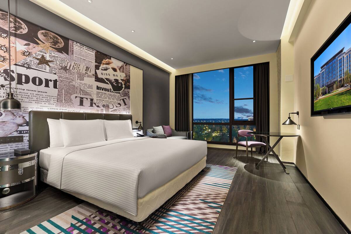 A modern hotel room with a large bed, a desk, a chair, and a TV.