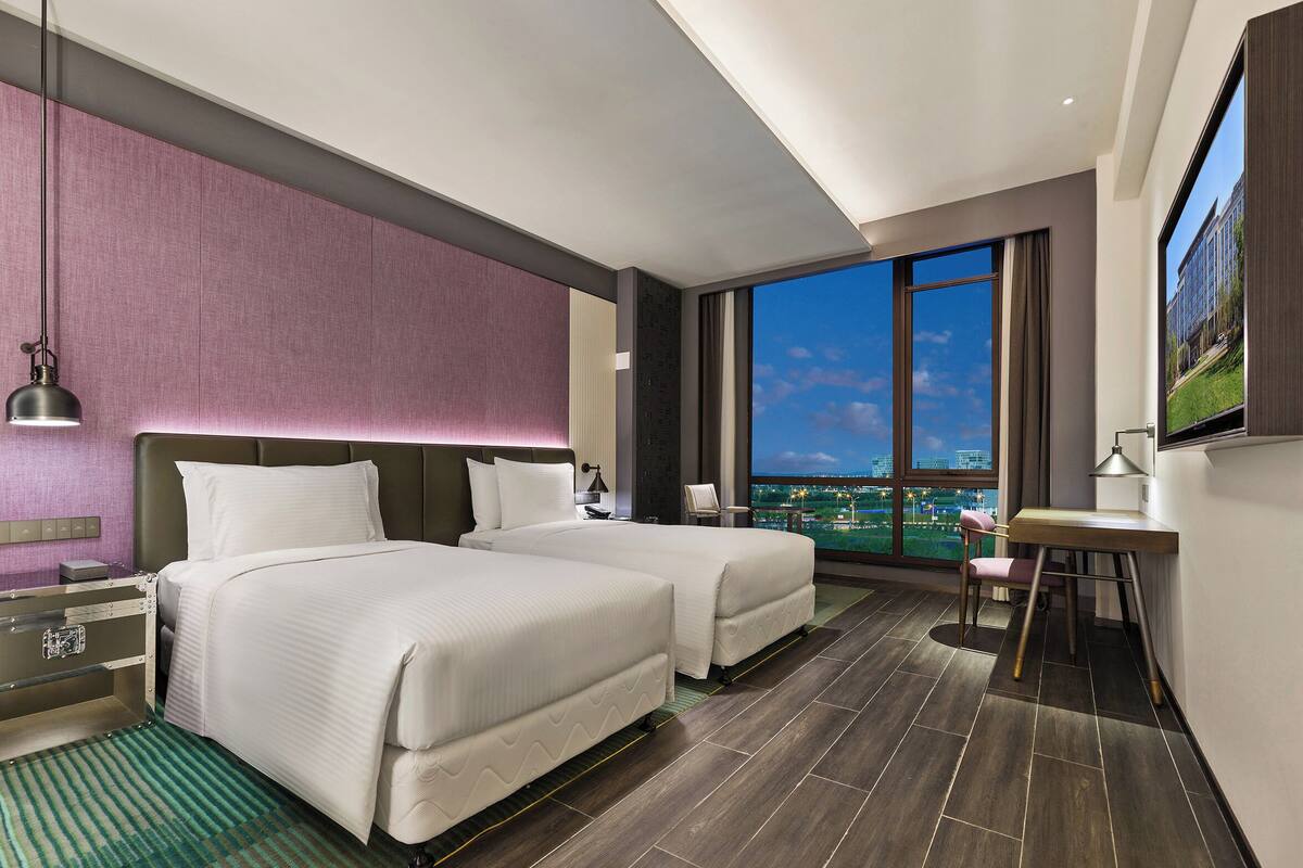 A hotel room with two beds, a desk, a chair, a TV, and a large window with a city view.