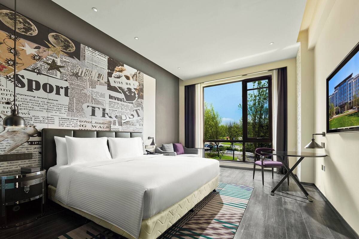 A modern hotel room with a large bed, a view of the outdoors, and a wall decorated with newspaper clippings.