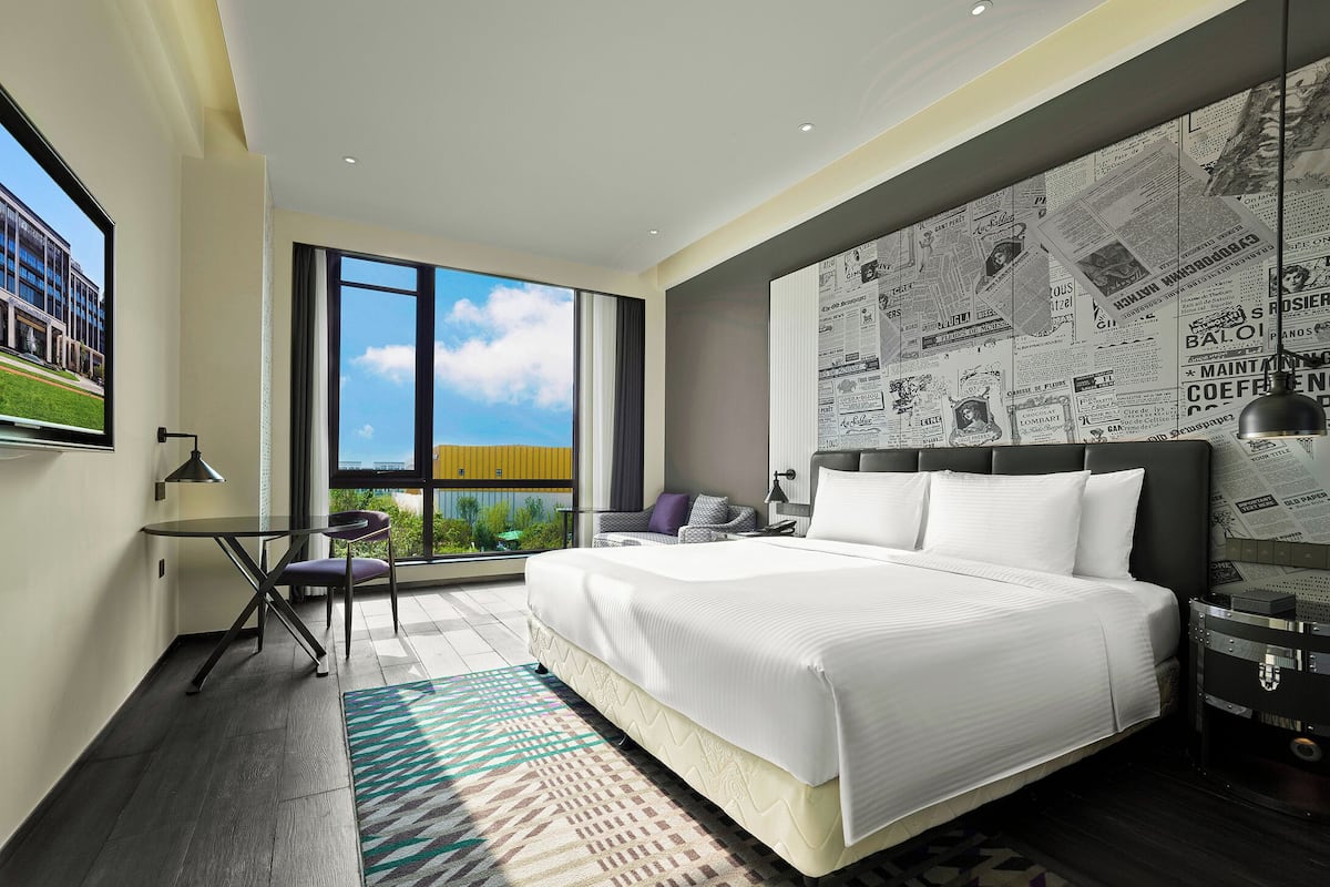 A modern hotel room with a large bed, a view of the outdoors, a desk with a chair, and a wall with newspaper clippings.