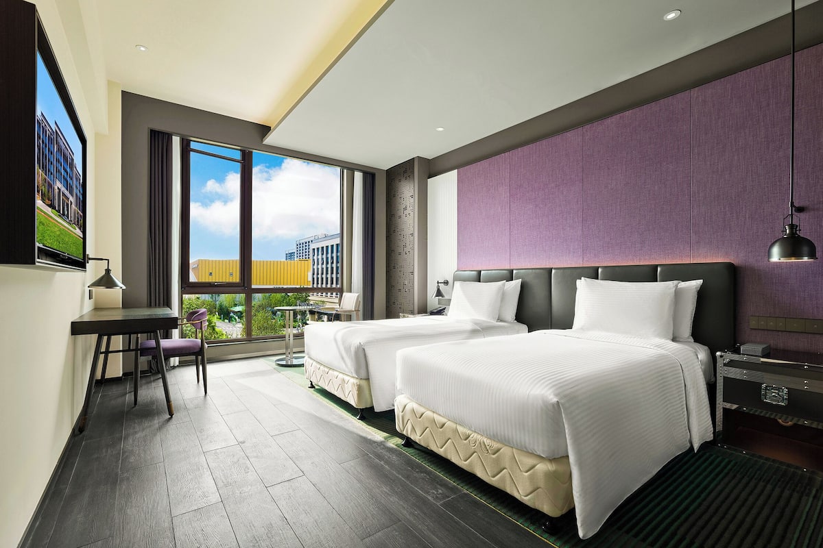 A modern hotel room with two beds, a desk, a chair, a large window with a view of buildings, and a ceiling light.