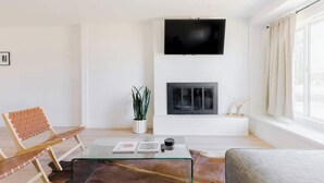A modern living room with a glass coffee table, a wooden chair, a sofa, a flat-screen TV mounted on the wall, a fireplace, and a large window with curtains. - Renovated Retreat, Old Town Scottsdale, Sleeps 6 (Scottsdale)