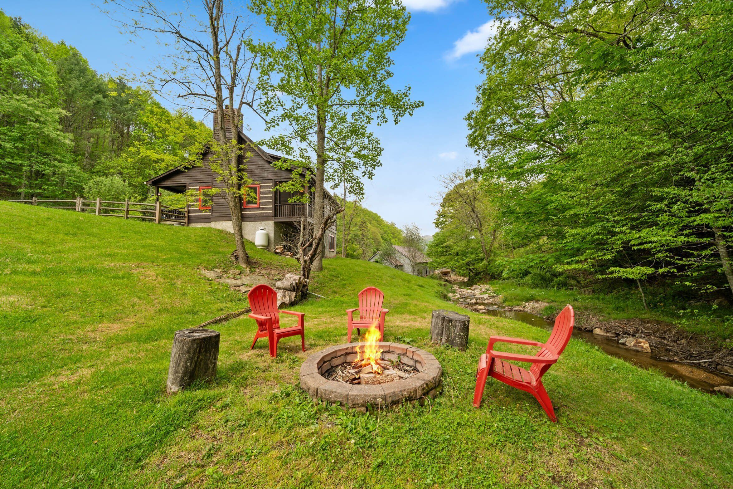A rustic cabin with a fire pit and red Adirondack chairs in a lush green setting.