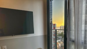 A room with a flat-screen TV mounted on the wall, a wooden console table with two small potted plants, and a large window with a view of buildings and a sunset. - Porto Privilege by VLV Stays (Salvador)