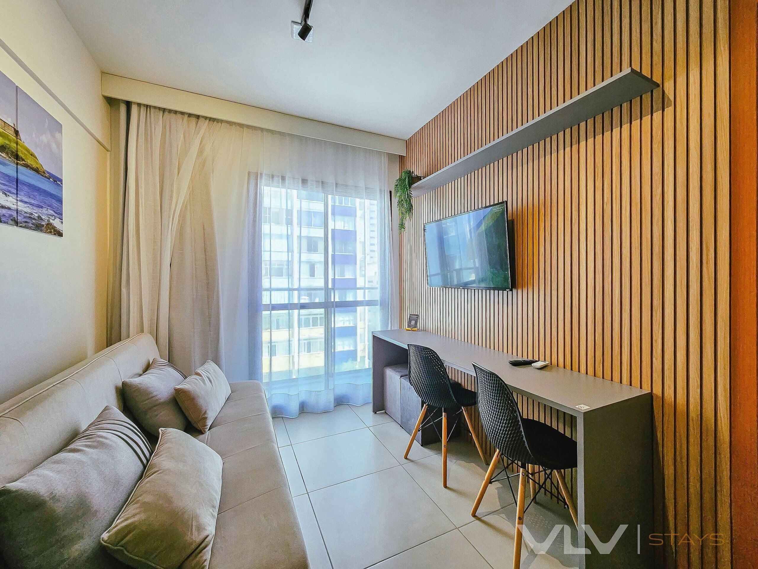A modern living room with a sofa, a dining area with chairs, a TV mounted on a wooden wall, and a large window with curtains.