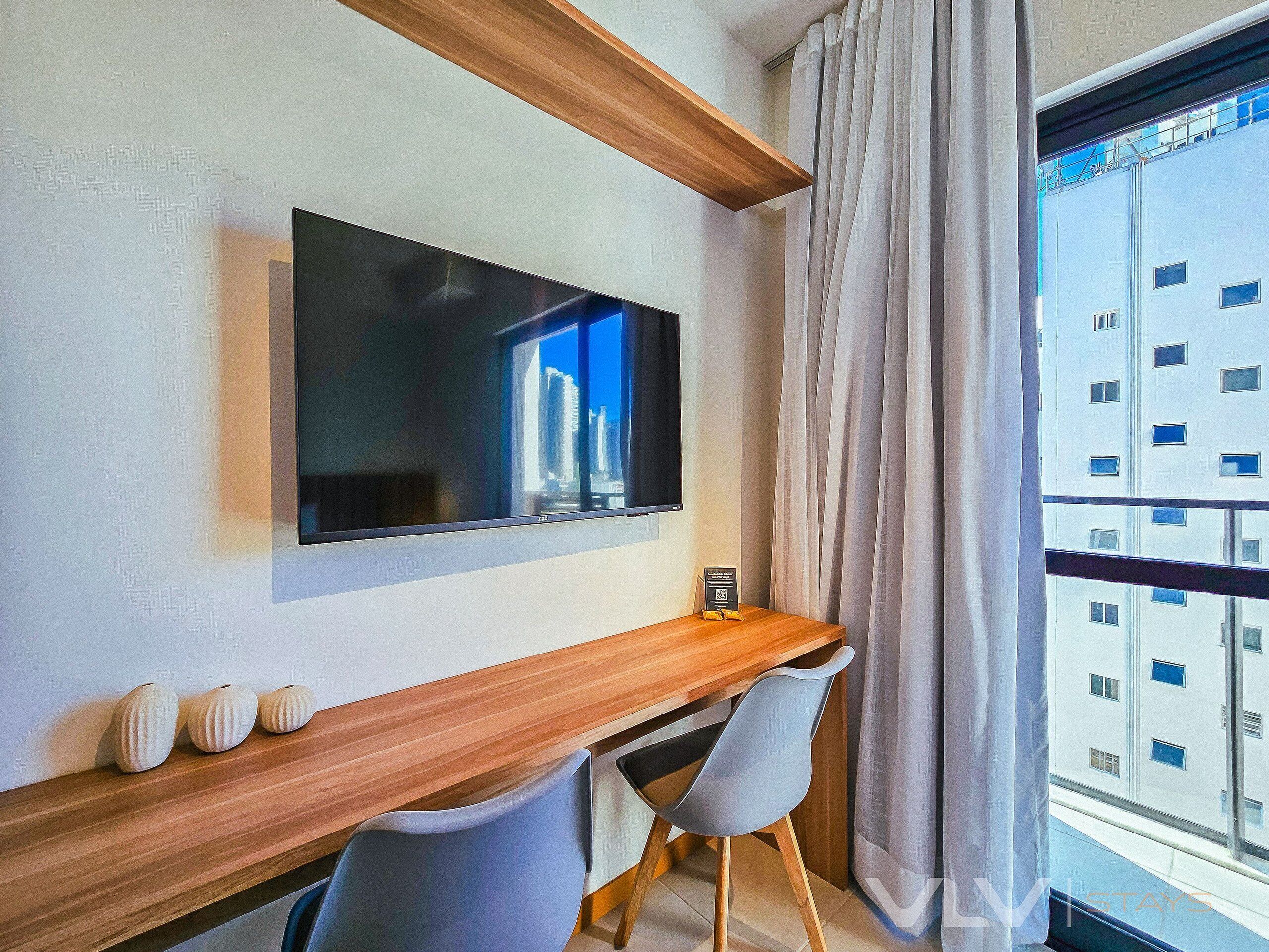 A modern hotel room with a large flat-screen TV mounted on the wall, a wooden desk, a grey chair, and a view of a building through the window.