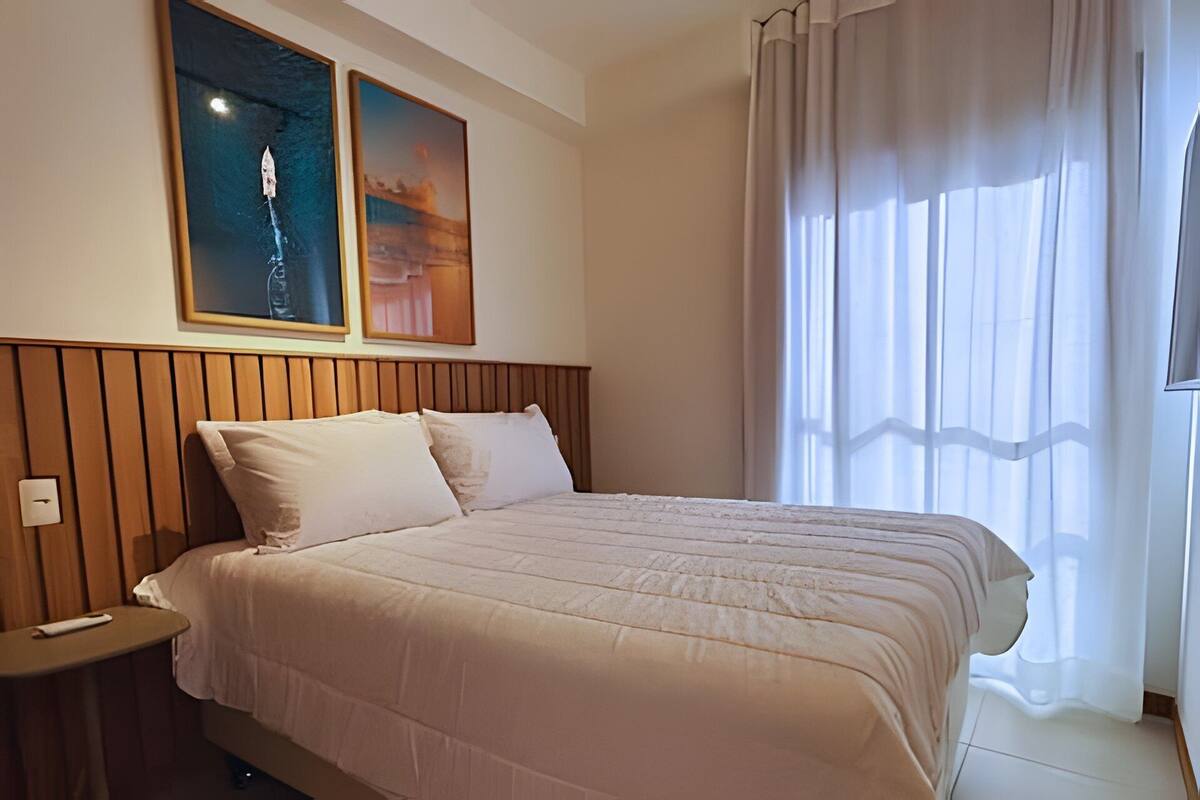 Una habitació d'hotel amb un panell de fusta al capçal del llit, un llit amb roba de llit blanca, dues obres d'art emmarcades a la paret i una finestra amb cortines transparents.