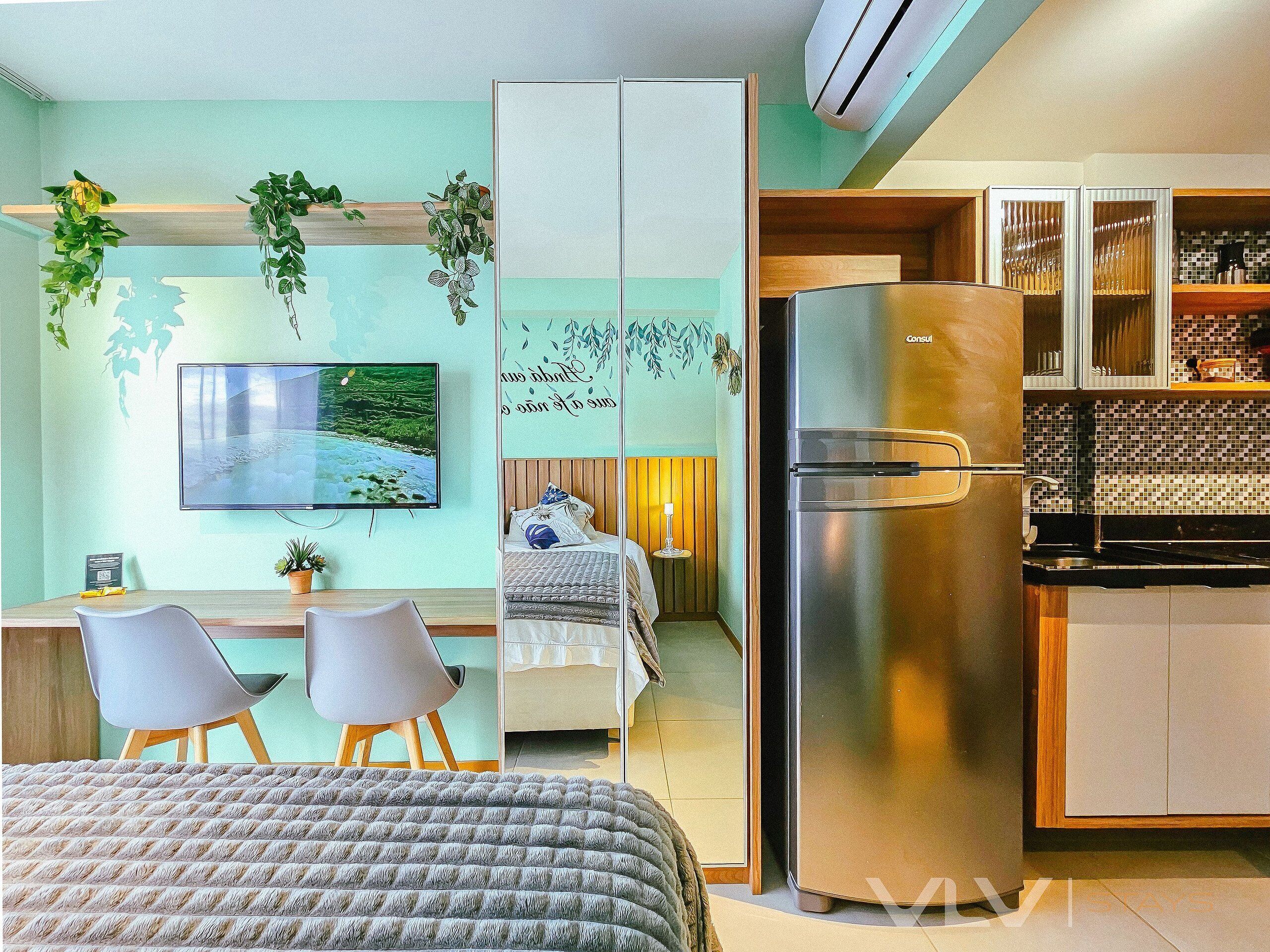 A modern hotel room with a bed, a desk with two chairs, a television, a refrigerator, and a kitchenette.