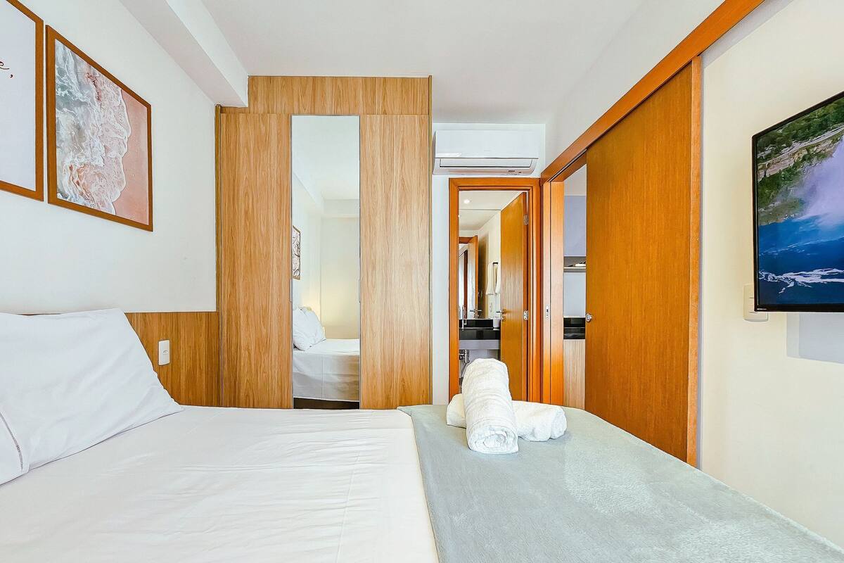 Habitació d'hotel amb un llit, una taula petita amb tovalloles, portes de fusta i una televisió penjada a la paret.
