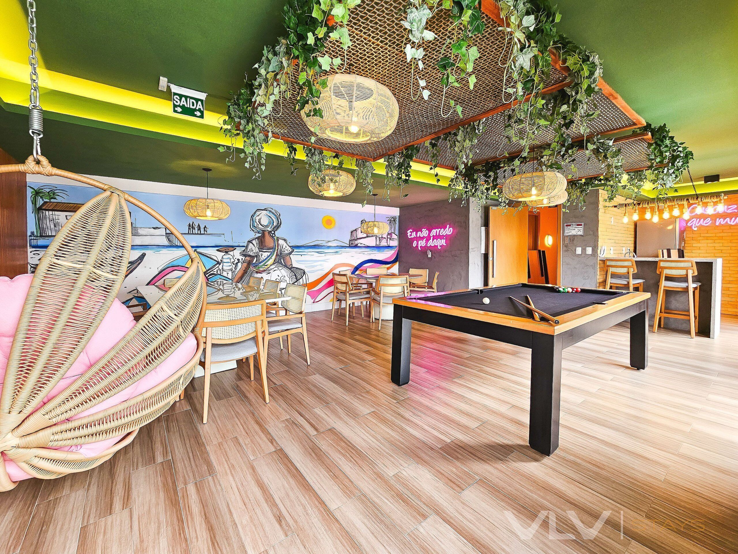 A pool table, chairs, a hanging chair, a mural, and a neon sign in a room with wooden flooring and green walls.