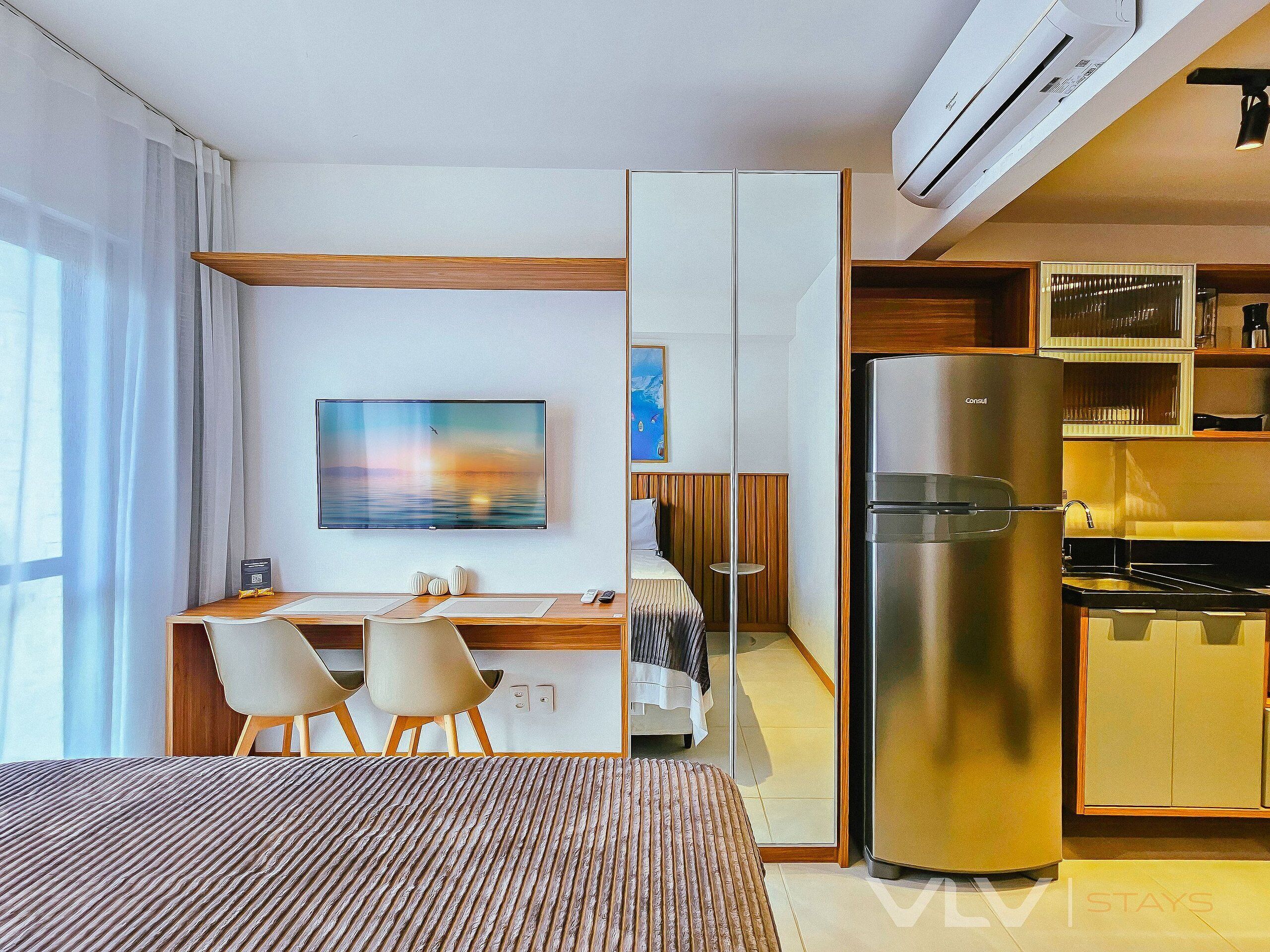 Quarto de hotel compacto com cama, escrivaninha com cadeiras, televisão, refrigerador e cozinha compacta.