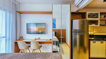 Quarto de hotel compacto com cama, escrivaninha com cadeiras, televisão, refrigerador e cozinha compacta.