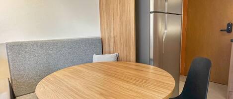 Una mesa pequeña y redonda con tapa de madera, una silla negra, un banco gris acolchado y un refrigerador de acero inoxidable.
