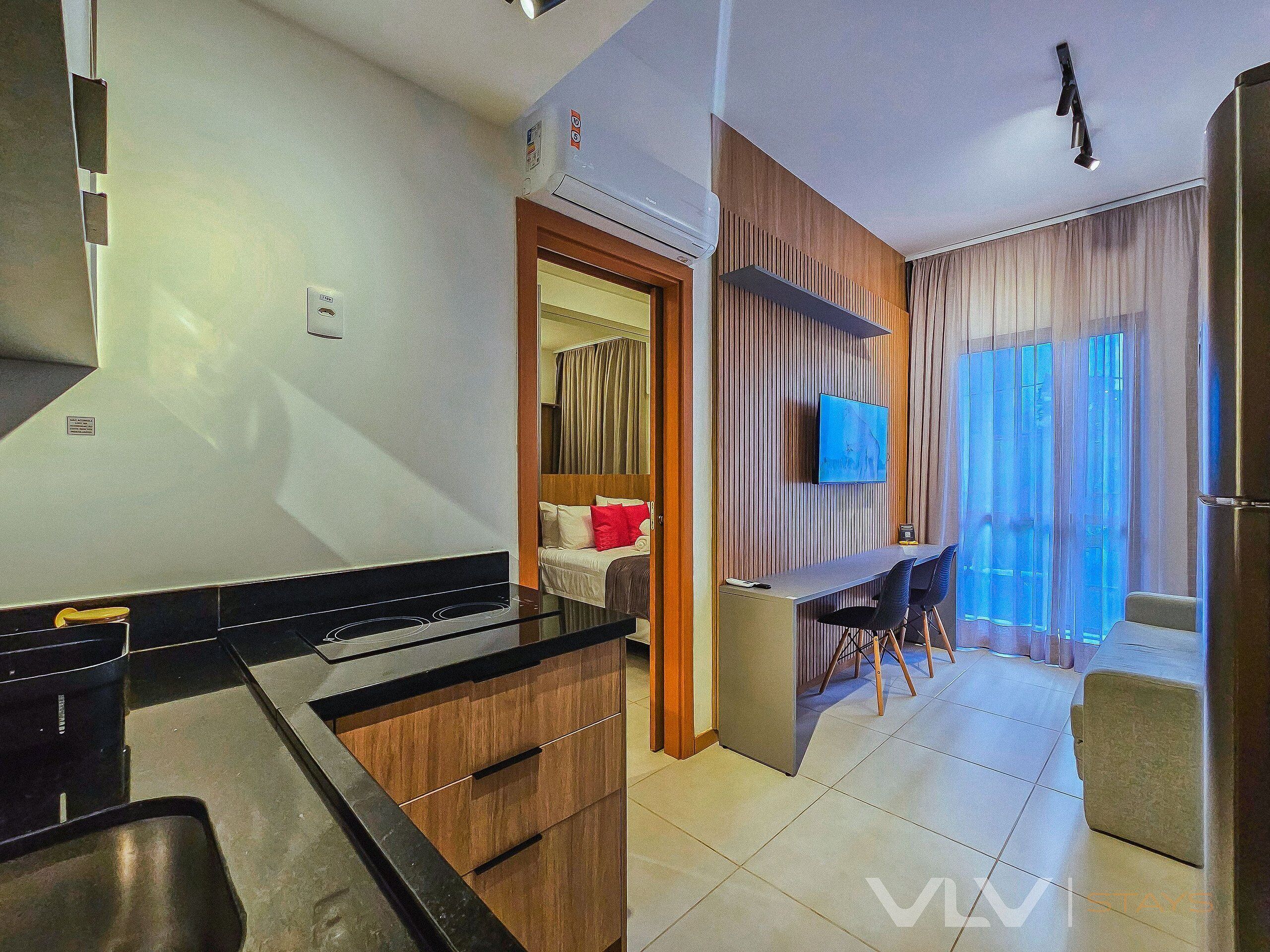 A modern kitchen with a stove, sink, and wooden cabinets, a dining area with a table and chairs, a TV mounted on the wall, and a sliding door leading to a balcony.
